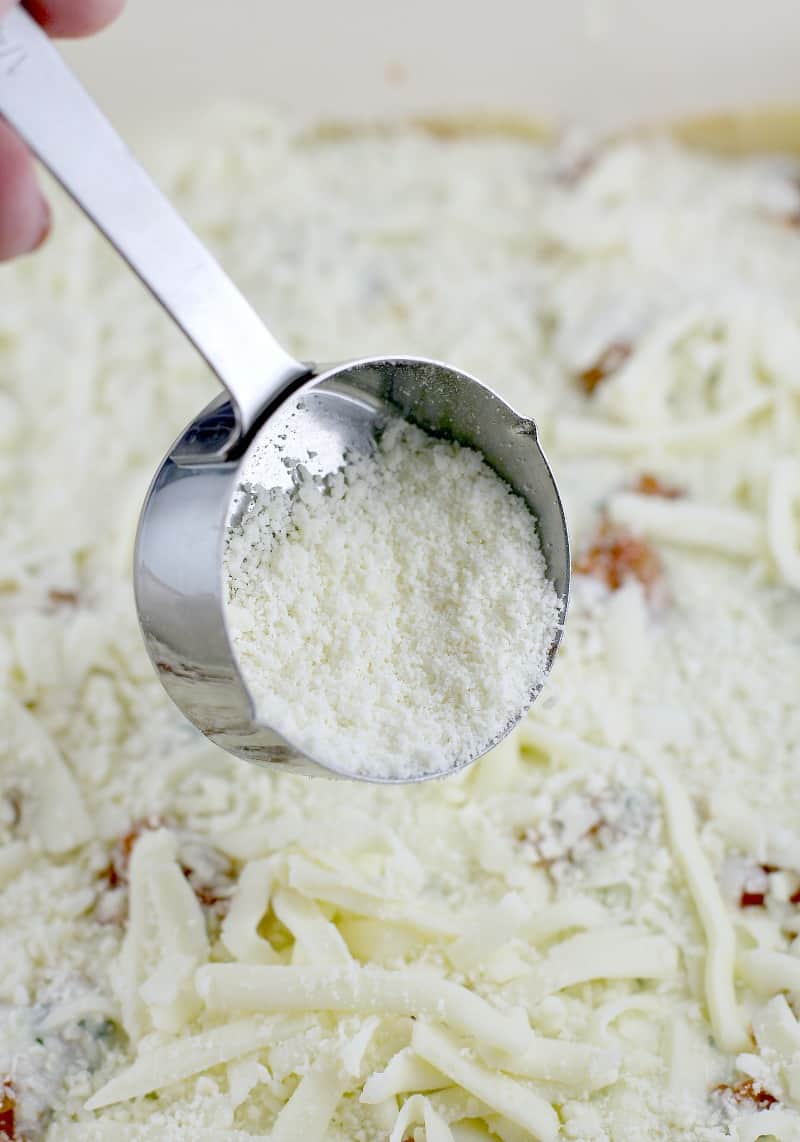a small metal measuring cup sprinkling grated parmesan over mozzarella in the lasagna dish
