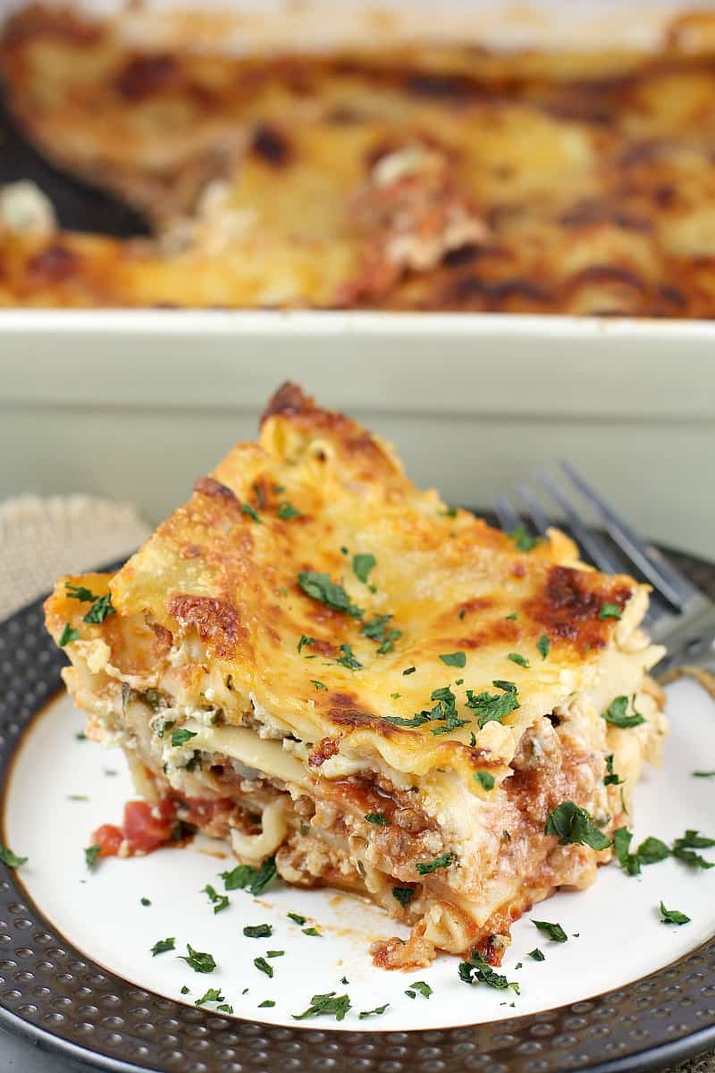 plated lasagna on a brown rimmed plate with the ceramic lasagna dish in the background