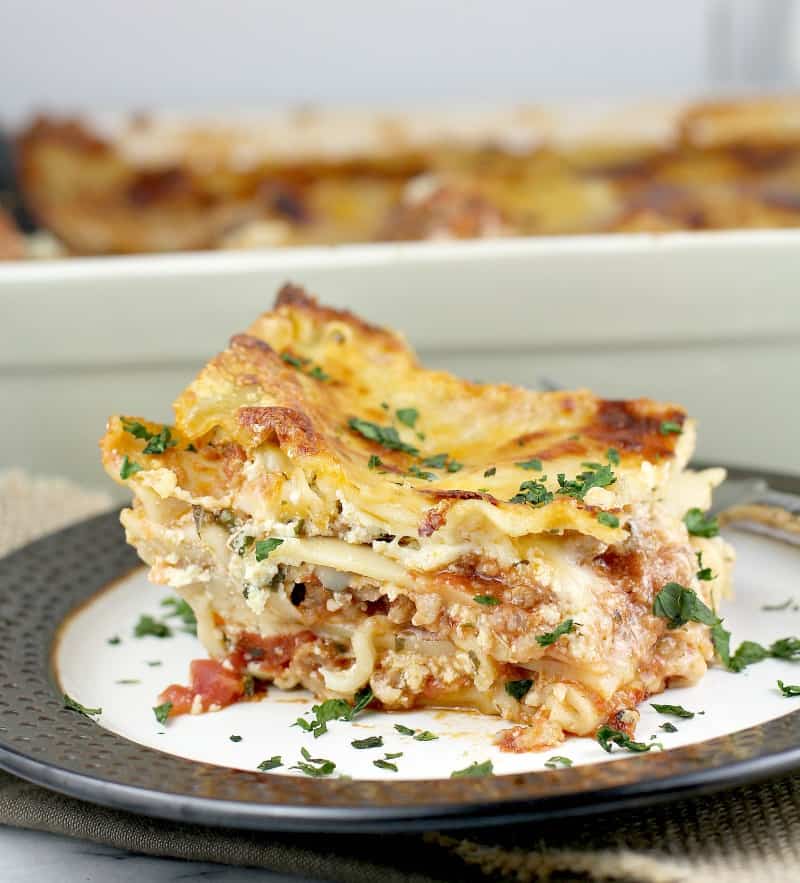 side view of a large square of lasagna plated on a white plate with brown rim, lasagna bake dish is in the back ground