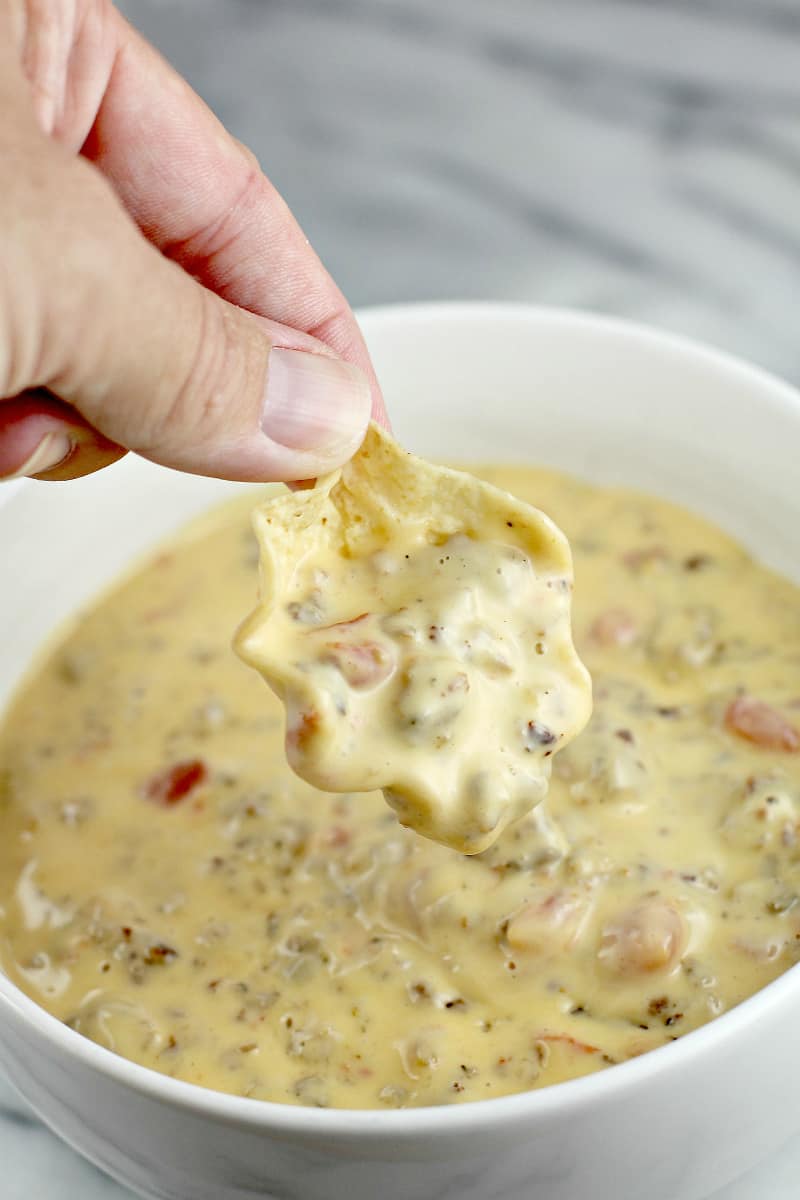 Hand holding a scoop tortilla chip with queso over a bowl full of queso