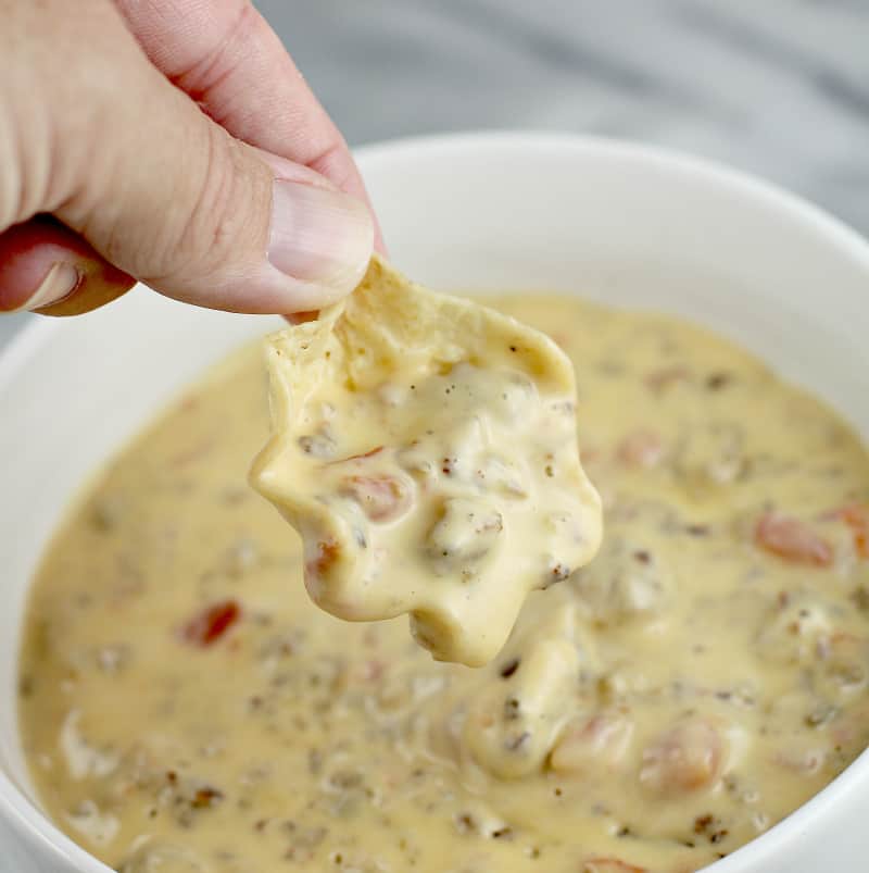 Close view of hand holding a scoop tortilla chip with queso over a bowl full of queso