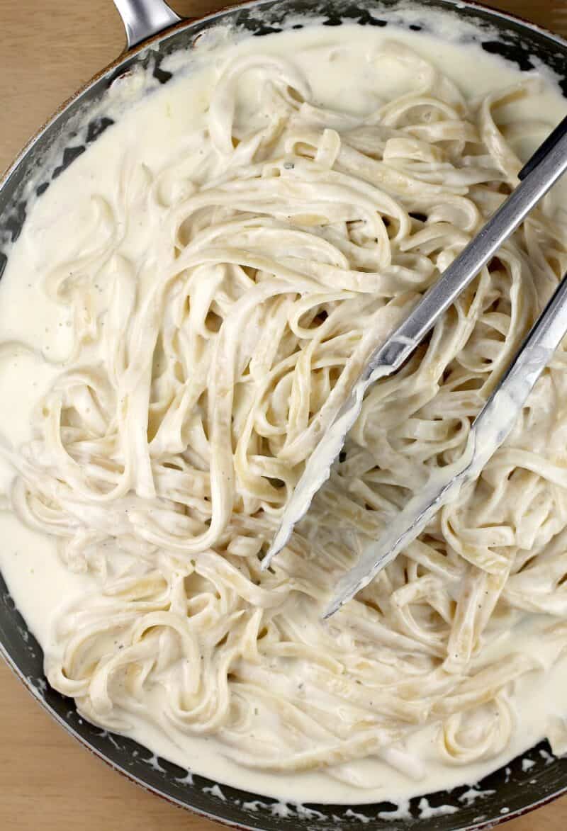 noodles added to the alfredo sauce with a pair of tongs