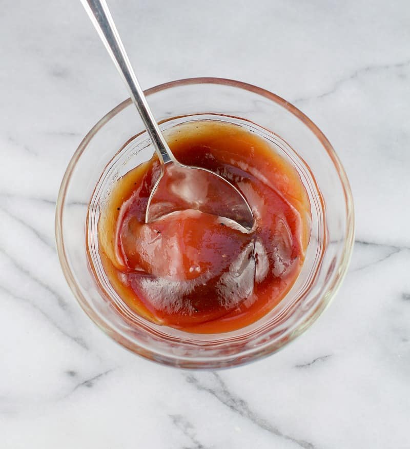top down view of a spoon mixing the ketchup and barbecue sauce in a clear glass bowl on a marble surface