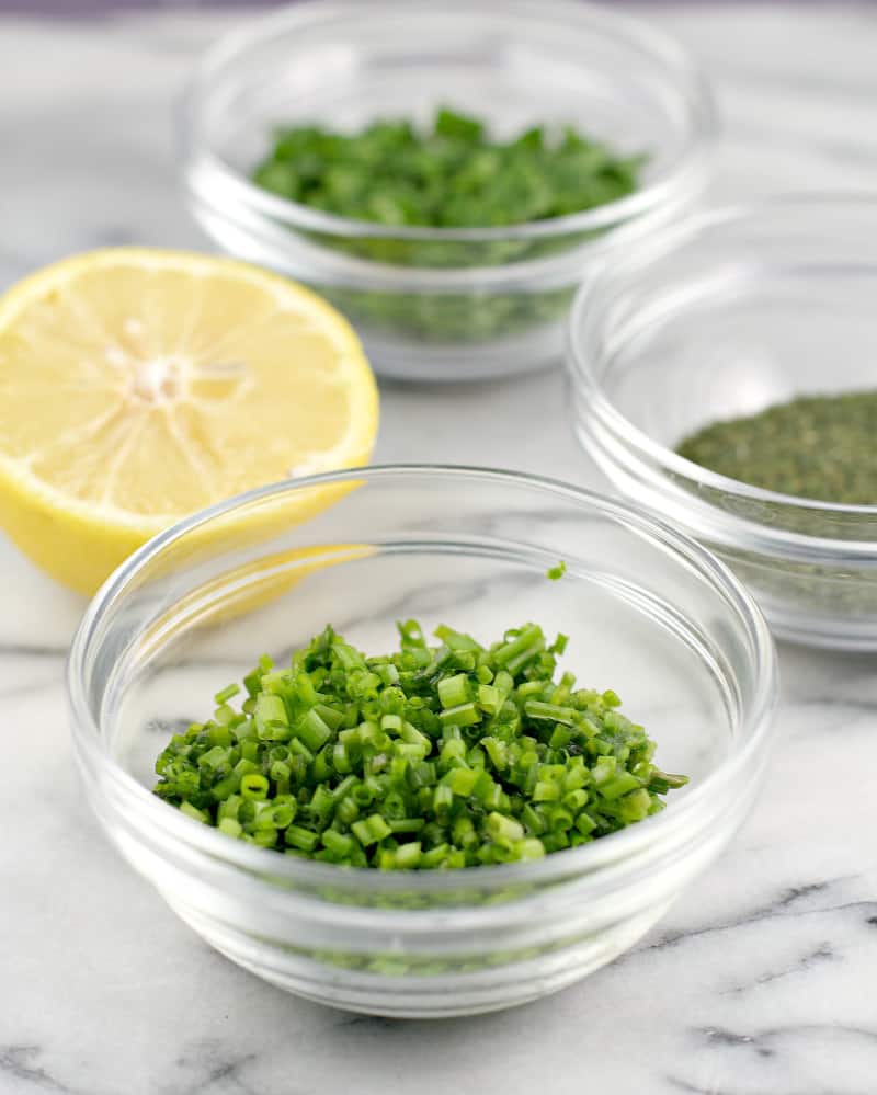 three small glass dishes showing the herbs parsley, chive, and dill, and a lemon half. marble background