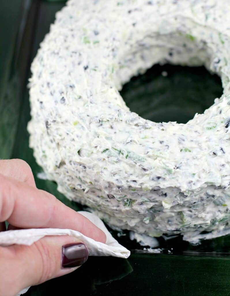 view of a hand holding a paper towel, removing smudges of dip from the plate after the christmas cheese wreath has been formed