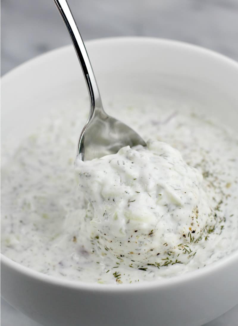 a silver spoon scooping healthy veggie dip tzatziki recipe in a white bowl, white marble background