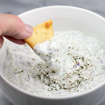 a hand scooping tzatziki sauce with a chip in a white bowl with marble background