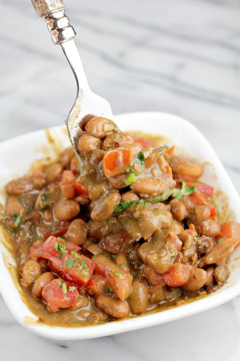 side view of the finished charro beans, in a small white dish on a marble surface. A fork is scooping beans up to show the ingredients and texture