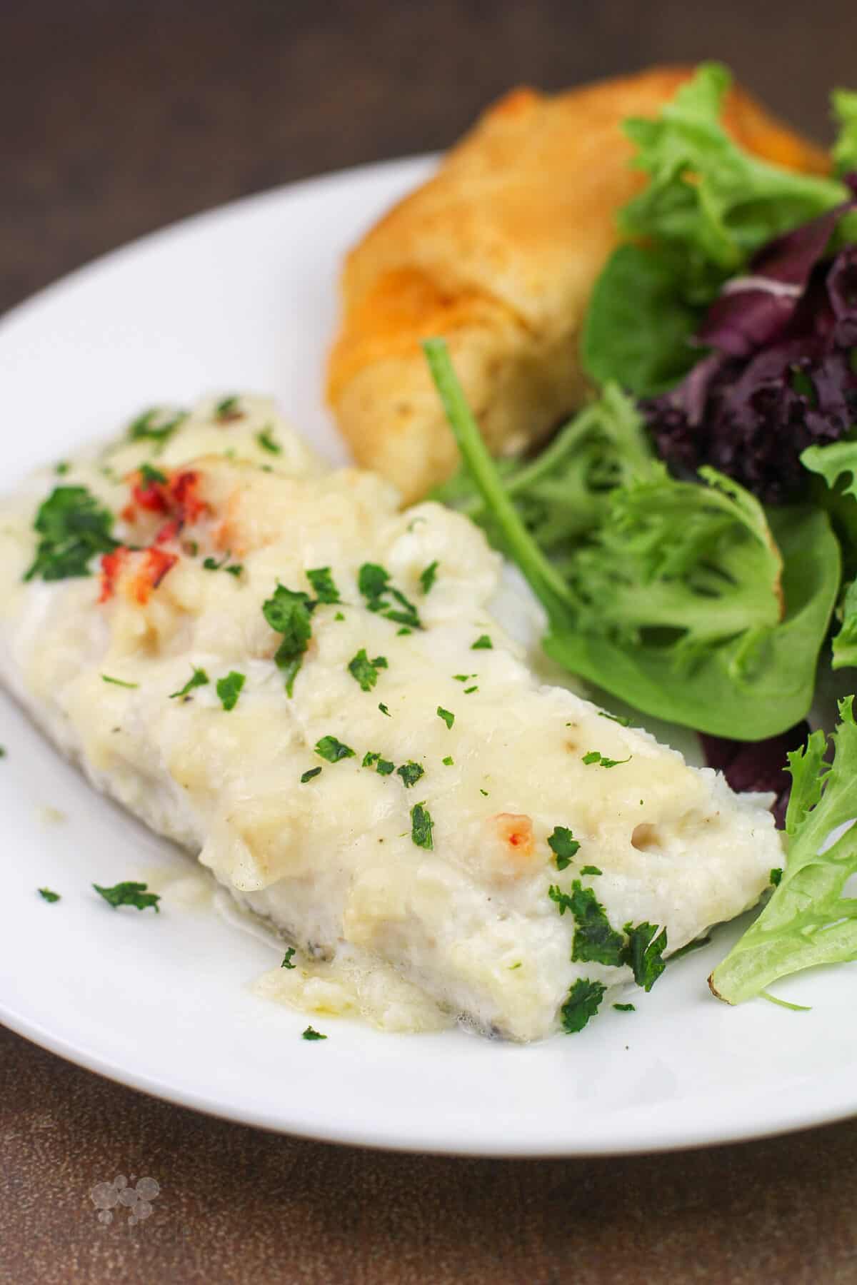portion of prepared haddock with lobster sauce on white plate with salad and roll