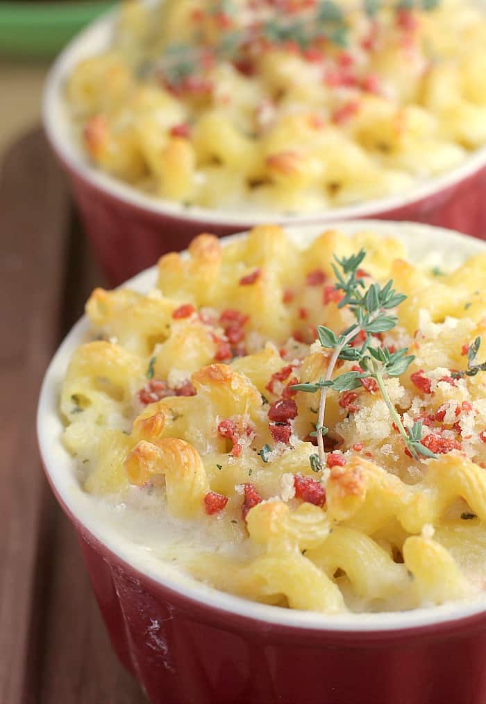 a red crock filled with macaroni and cheese topped with bacon and a tiny sprig of thyme; a second crock is in the background