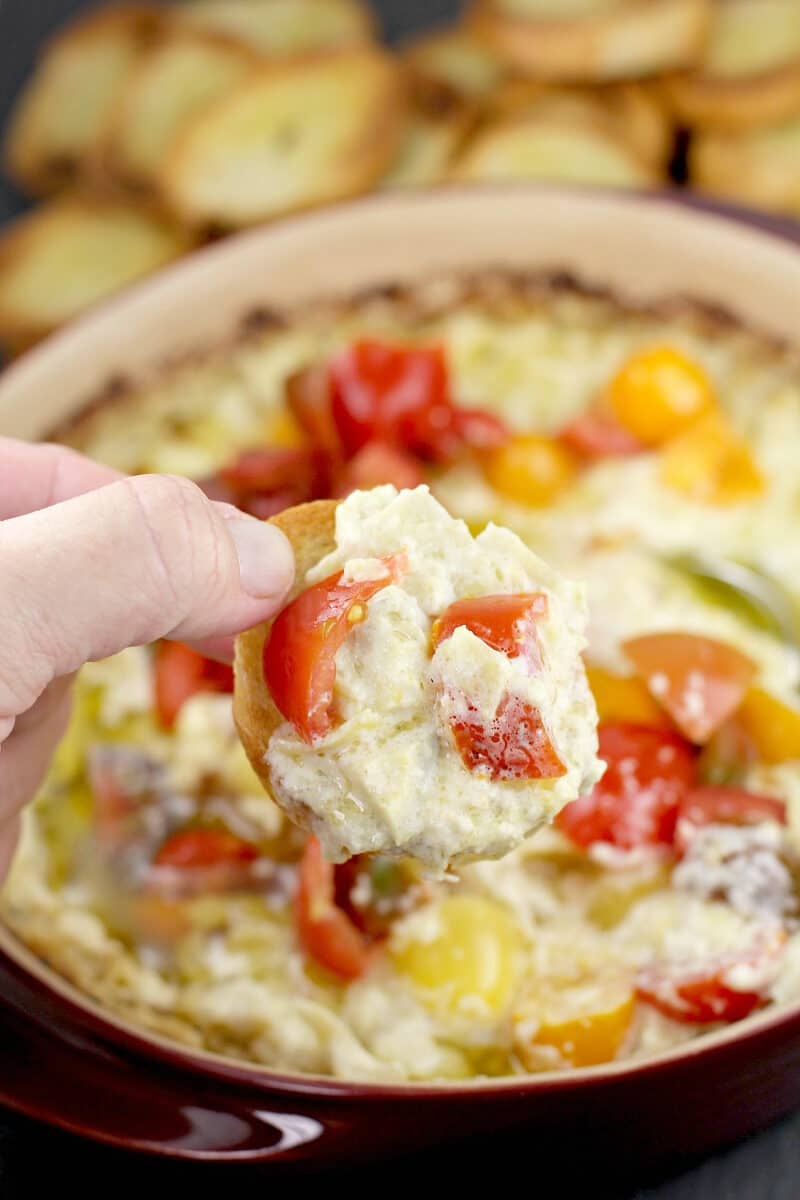 hand holding a piece of toasted bread with green chili artichoke dip