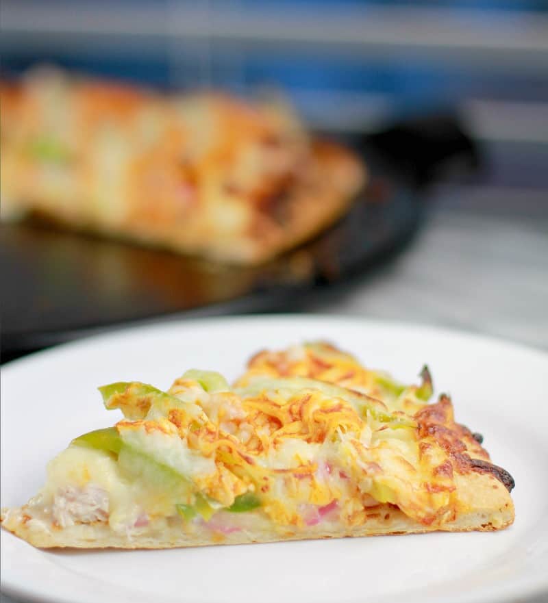 Side view of a slice of pizza on a white plate (green bell pepper, turkey, and onion toppings are peeking out from the cheese); finished pizza is in the background on a black pizza stone