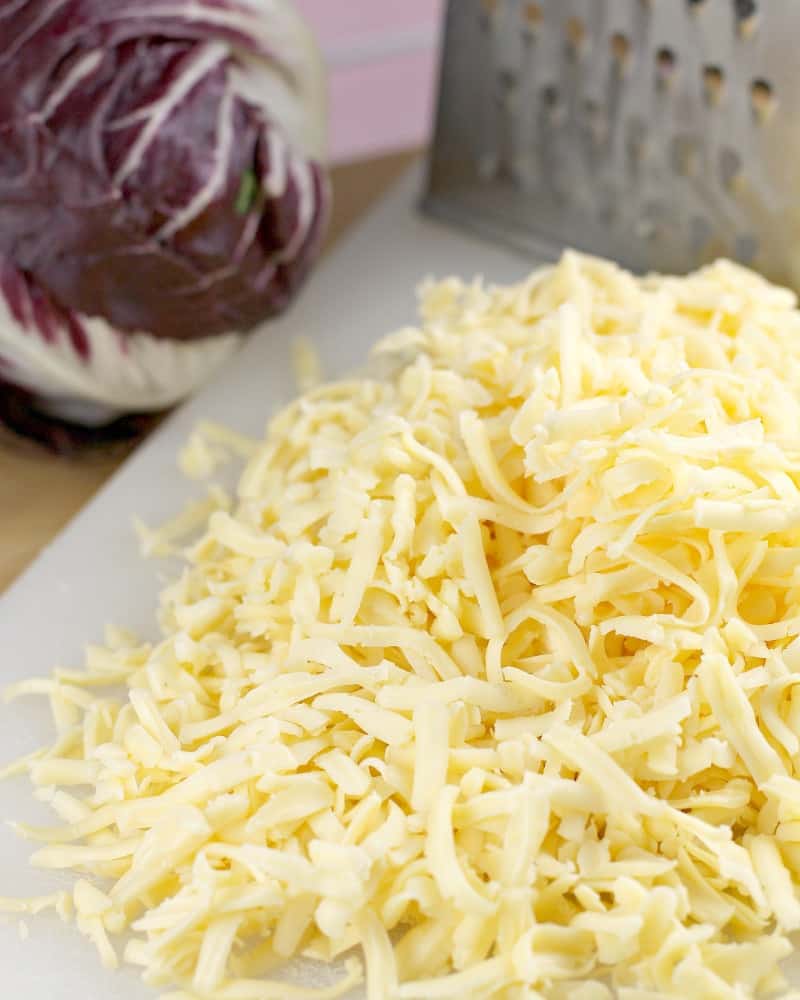 a pile of shredded Havarti cheese on a white cutting board cheese grater and raddichio are in the background to show the ingredients for this easy appetizer