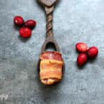 bacon wrapped bite on wood spoon, cranberries sprinkled around; blue background