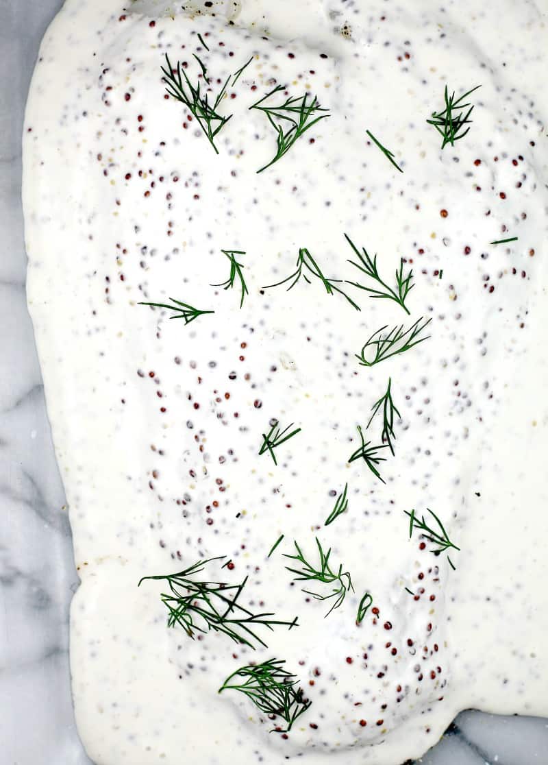 cod laying in a bake dish, topped with the mustard cream, with fresh dill fronds sprinkled over top, ready for the oven