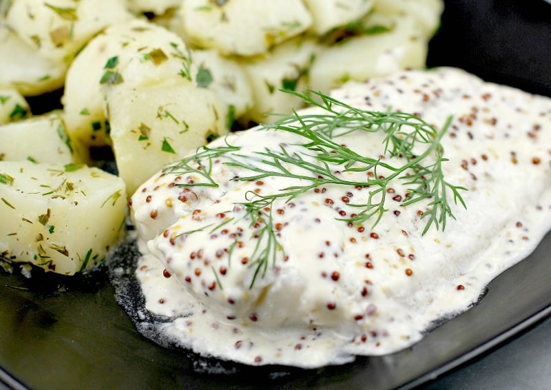 cropped view, finished baked fish on a black dish on a marble surface. Herbed potatoes are in the background