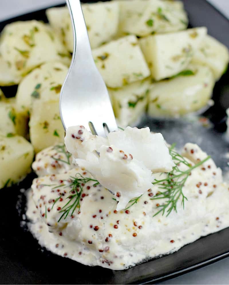 finished baked fish on a black dish on a marble surface with a fork lifting up some fish to show texture. Herbed potatoes are in the background