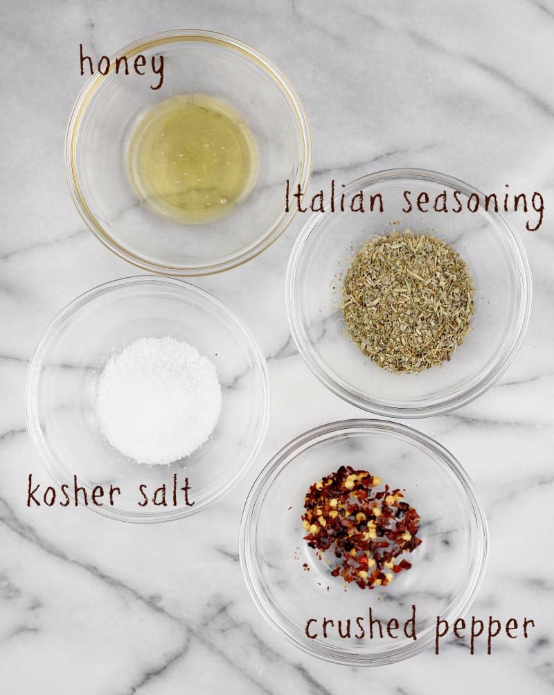 marble board on which are 4 small glass bowls showing ingredients: honey, italian seasoning, salt, and crushed red pepper