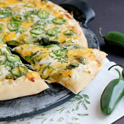 side view of the finished pizza on a black pizza stone with one slice cut and nudged out a bit to show melty cheese. a white napkin with green stencil and fresh jalapenos are placed as decoration