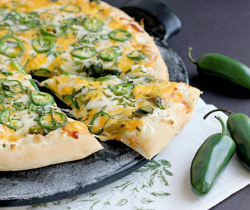 side view of the finished jalapeno pizza on a black pizza stone with one slice cut and nudged out a bit to show melty cheese. a white napkin with green stencil and fresh jalapenos are placed as decoration