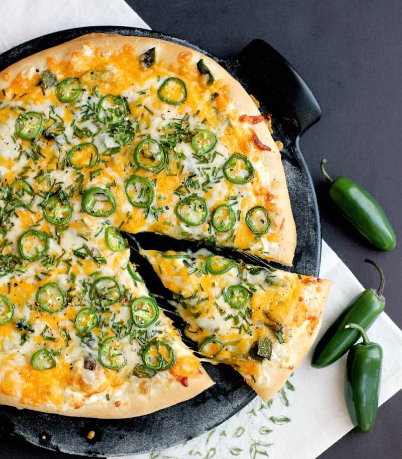 top view of the finished jalapeno popper pizza on a black pizza stone with a black background. a single slice is cut in the pizza and pulled out a bit.