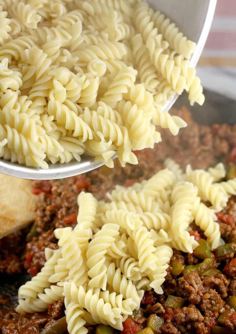 cooked pasta being poured into a skillet with the rest of the ingredients