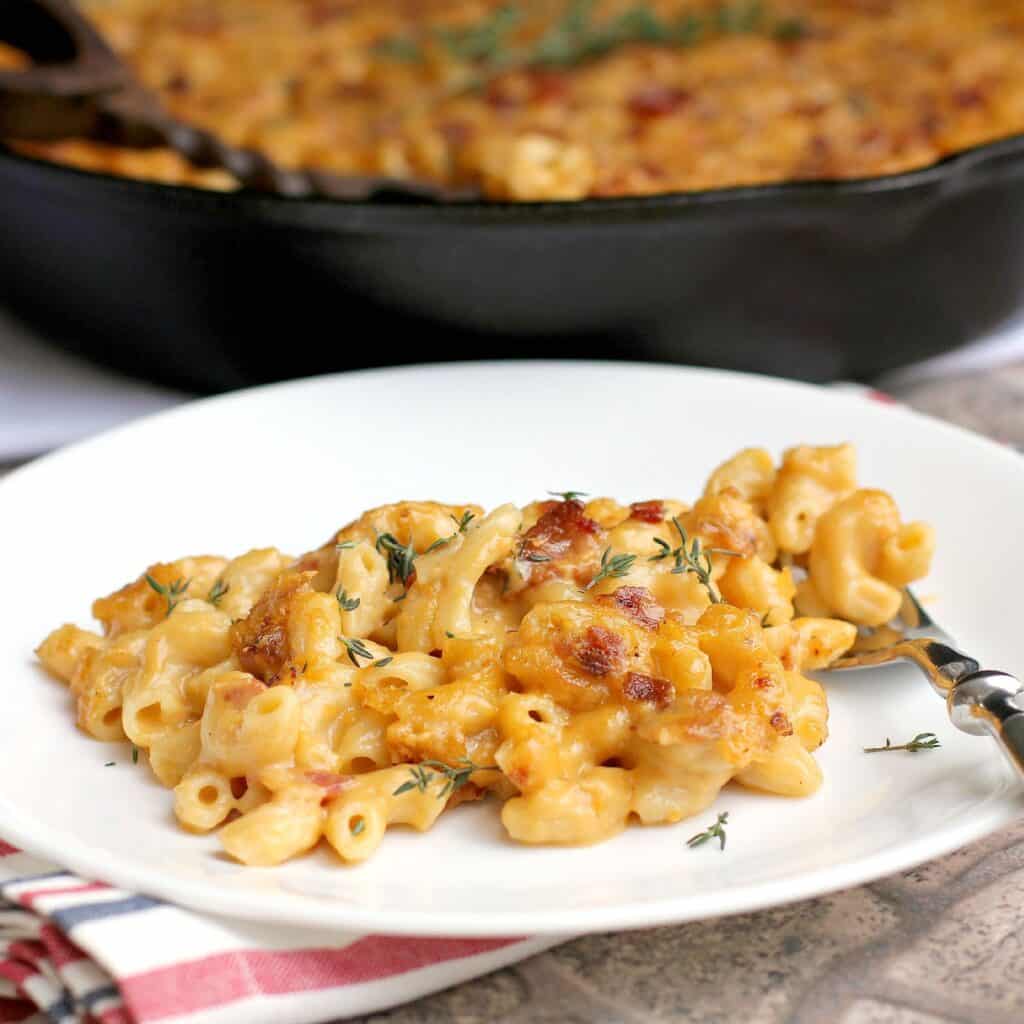 side view of finished bacon macaroni and cheese plated on a white dish; remainder of the mac n cheese is in the background in a cast iron skillet (zoomed view)