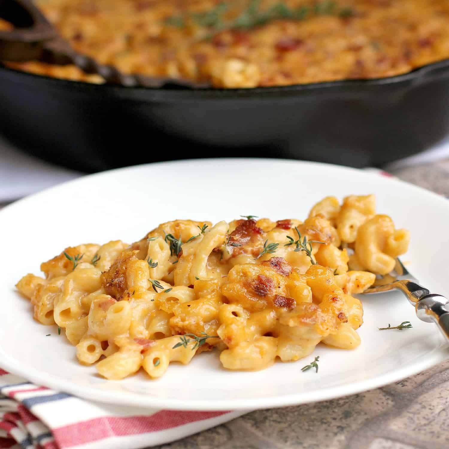 side view of finished bacon macaroni and cheese plated on a white dish; remainder of the mac n cheese is in the background in a cast iron skillet (zoomed view)