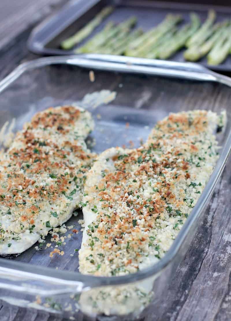 view of cooked parmesan crusted haddock in clear bake dish on wood surface; asparagus in background