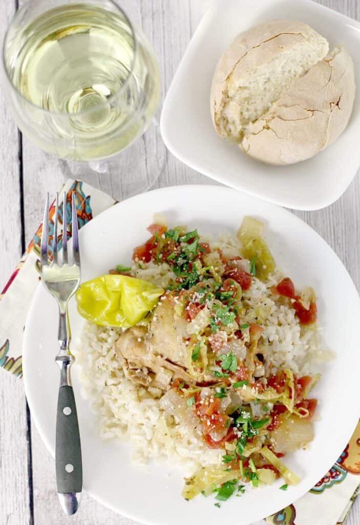 Top down view showing cooked slow cooker italian pepperoncini chicken served over rice on a white dish with a fork on the side. Bread and glass of white wine are at the top.