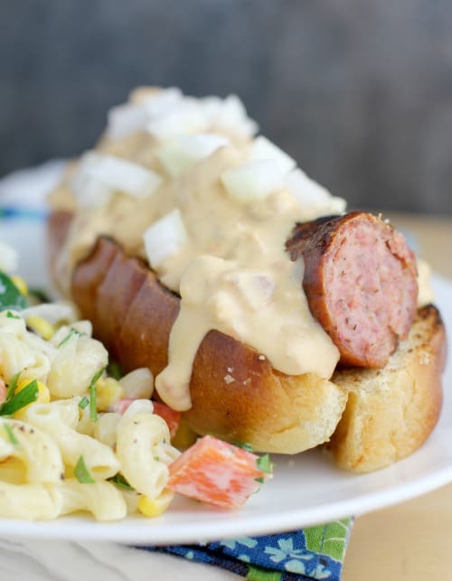 similar view of a finished beer kielbasa with queso, this time in a hot dog bun, white plate, with potato salad on the side