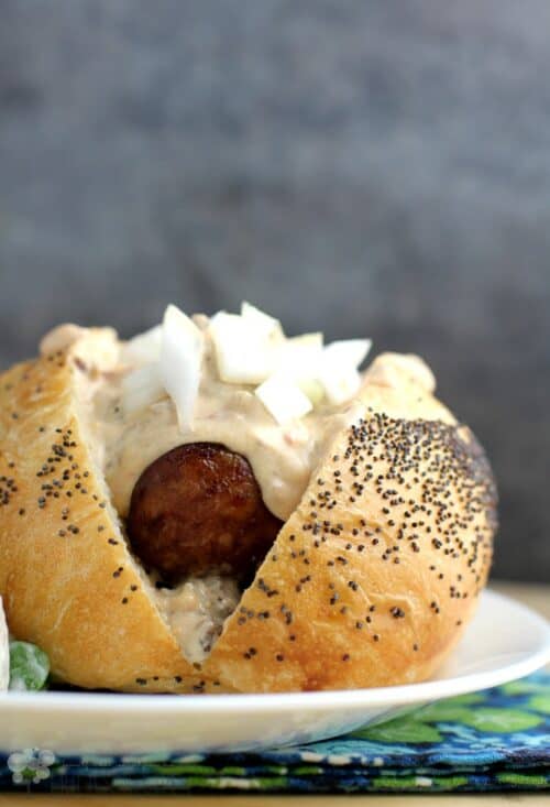 front view of the assembled beer brat in a bun with Guinness extra stout queso on top; on a white plate with a grey-blue background