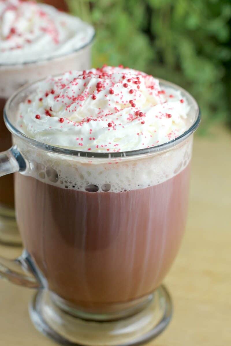 red velvet latte in a glass mug on a wood board; second cup and green leaves of a plant in the background