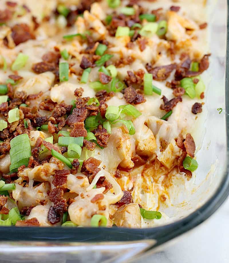 view showing the bacon and cheese on the rest of the dip in a clear glass bake dish, with a corner scooped out to show the under layers of chicken and cream cheese