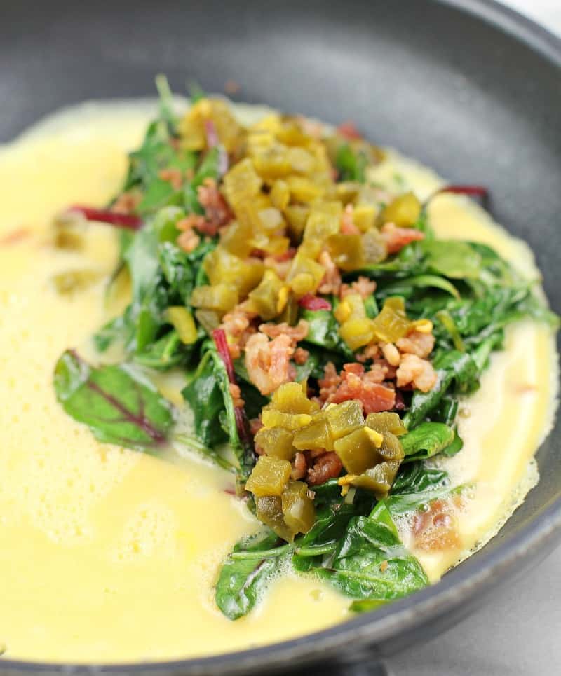 Side view of omelet toppings (greens, bacon, and jalapeno) on egg cooking in skillet.