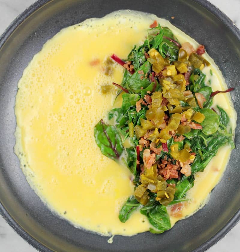 Top down view of omelet toppings (greens, bacon, and jalapeno) on egg cooking in skillet.