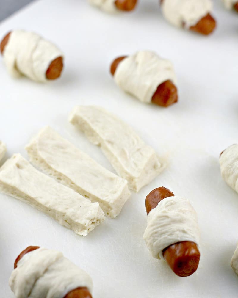 strips of pizza dough with several wrapped mini pigs in blankets on a white board