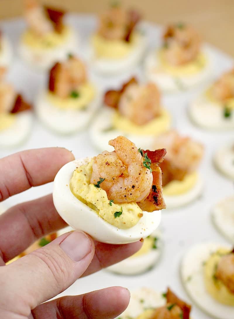 a hand holding up one deviled egg with bacon and shrimp; a white platter of others is in the background