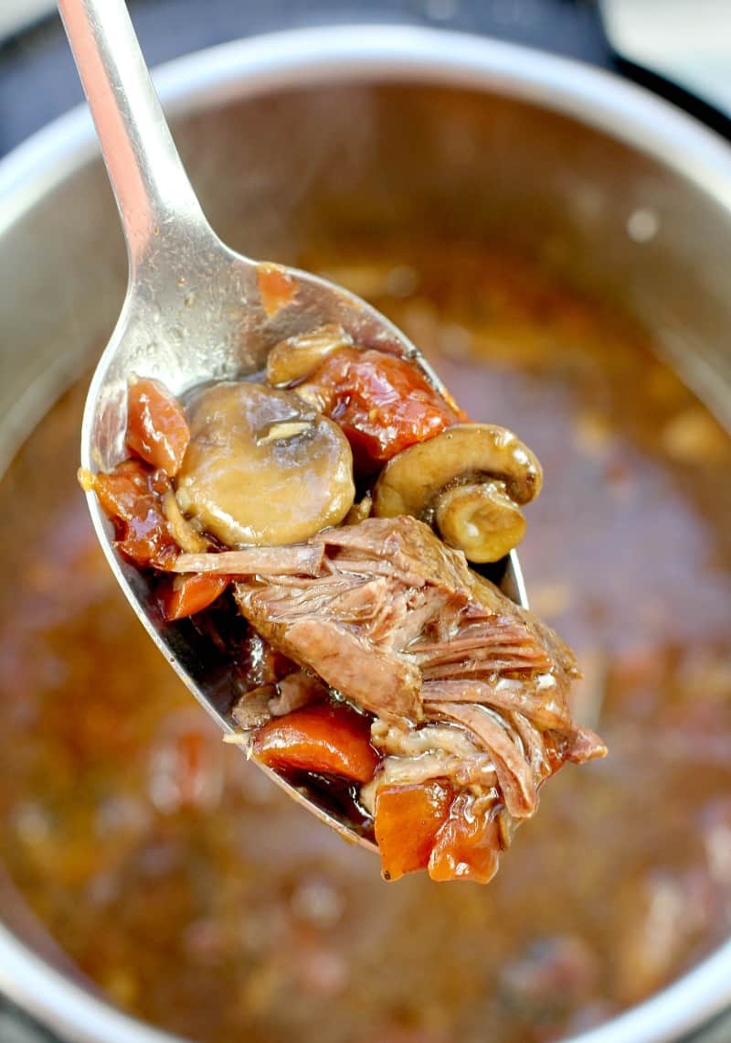 a large serving spoon scooping up cooked pot roast with beer, rest of the pot and recipe are in the background