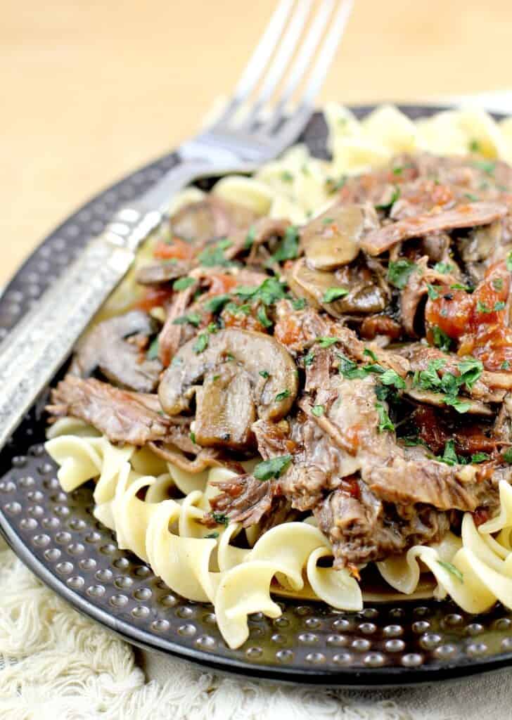 side view from the left of the pot roast with beer over noodles on a plate with a fork, placed on a cutting board