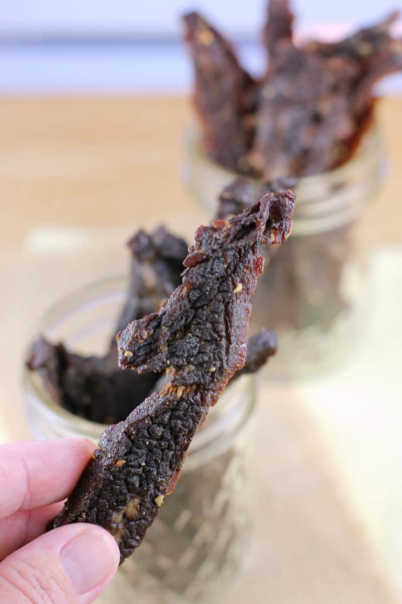 hand holding a piece of jerky; mason jars filled with jerky in the background