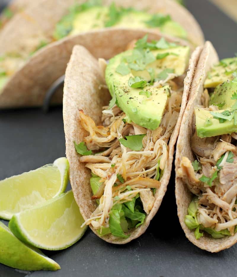 2 whole wheat tortillas stuffed with lettuce, chicken, and avocado on a slate with lime wedges