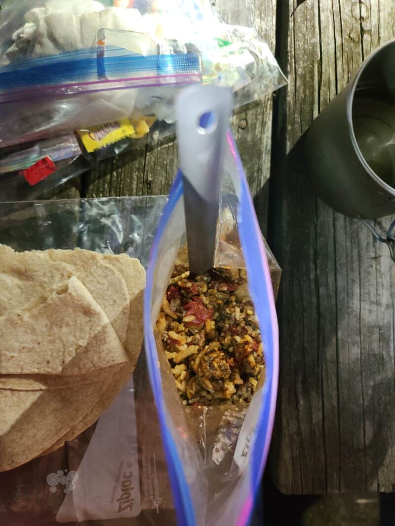 picnic table surface with tortilla and ziploc bag with backcountry sloppy joes and spoon; metal mug off to the side 