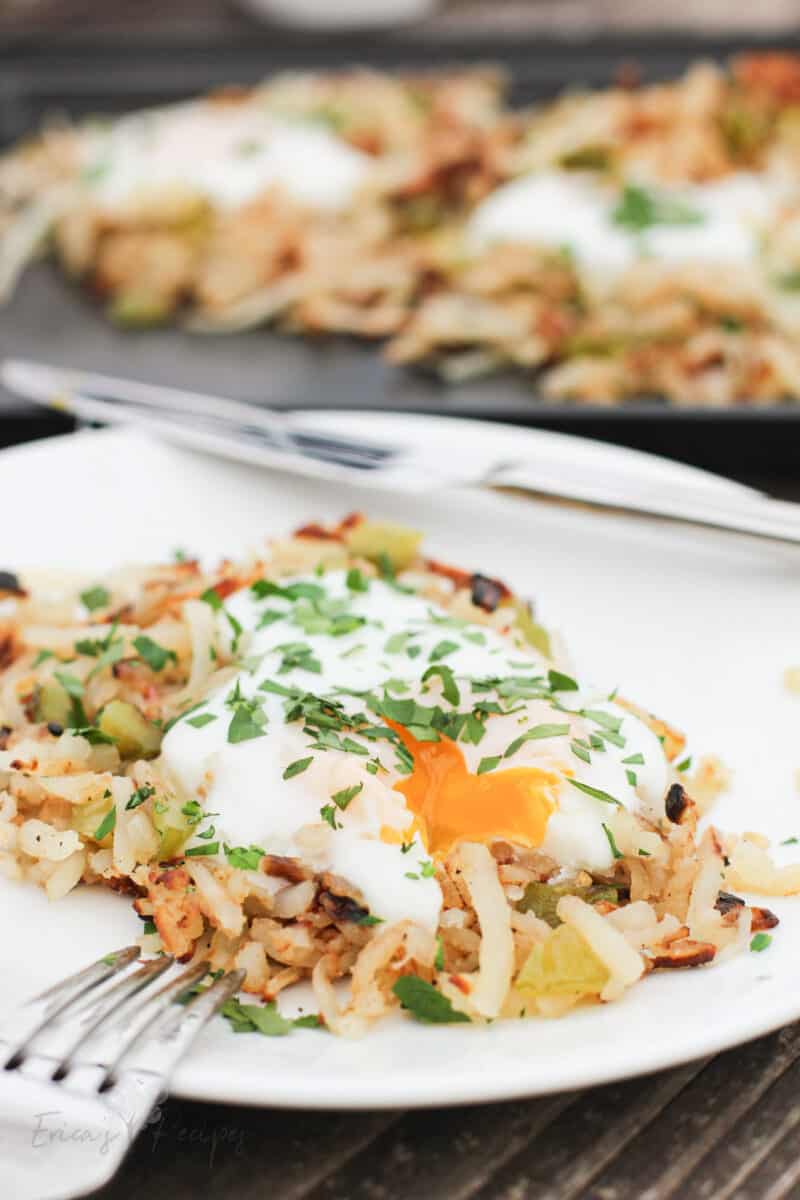 serving of griddle hash browns on white plate with egg broken and yolk running out onto potatoes; rest of potatoes on sheet in background