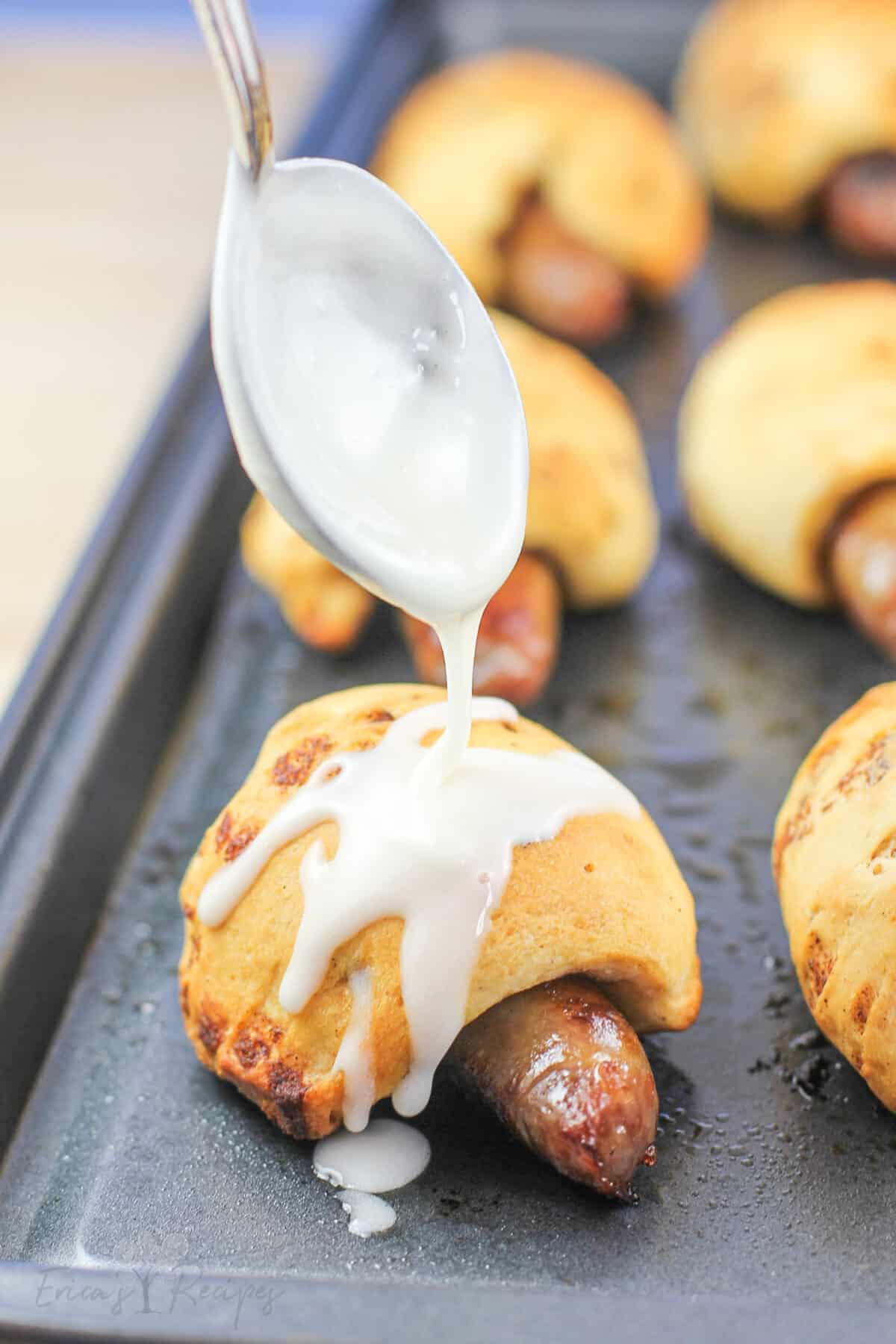 cinnamon roll pigs in a blanket on metal bake sheet; spoon is drizzling icing over top